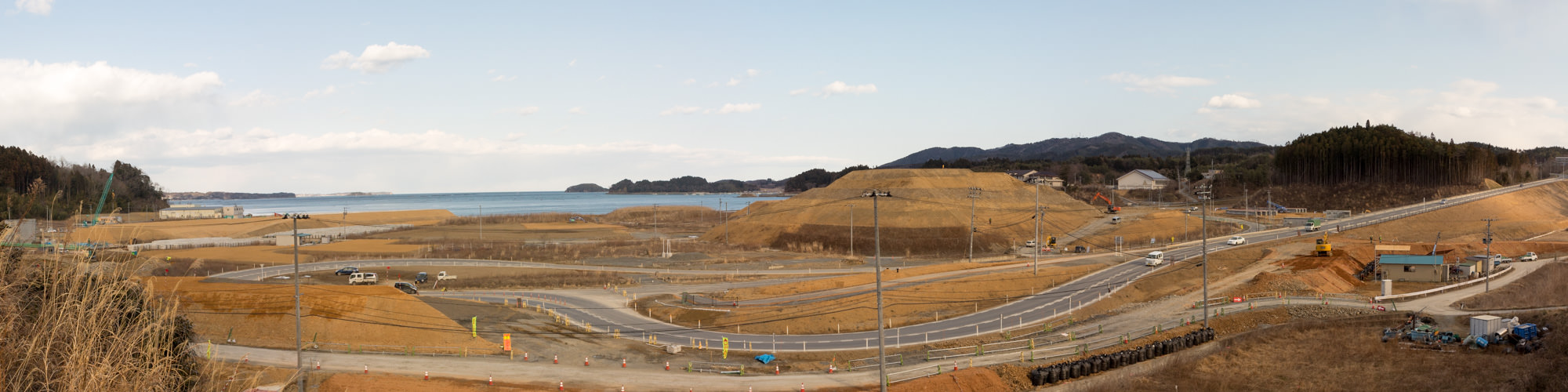 手前には複数の舗装された道路があり、車両が走行し道路の周囲には工事用のコーンやバリケードが設置され、安全対策が施されている海岸沿いに広がる大規模な建設現場の様子の写真