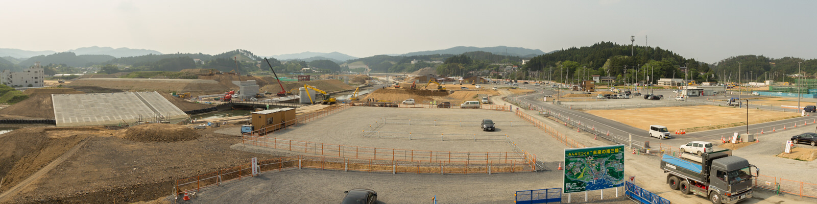 広い工事現場にクレーンやトラックなどの重機が点在し、舗装が始まった区画もある中、周囲には木々と丘が広がり、右下には案内板が設置されている工事現場の様子のパノラマ写真