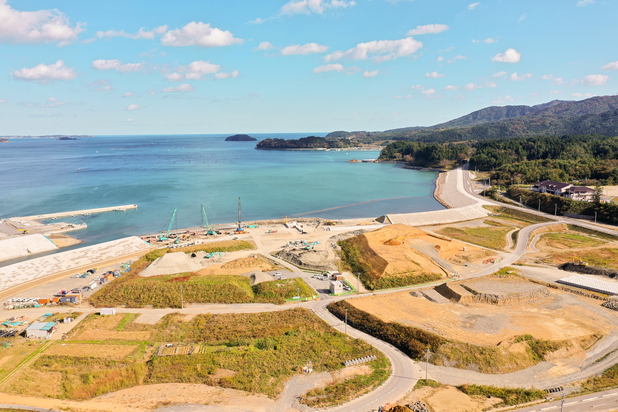 澄んだ青い海に小島が点在し、岸辺には大型クレーンが工事を行っており、周囲には重機や土砂が配置されている湾岸開発の写真