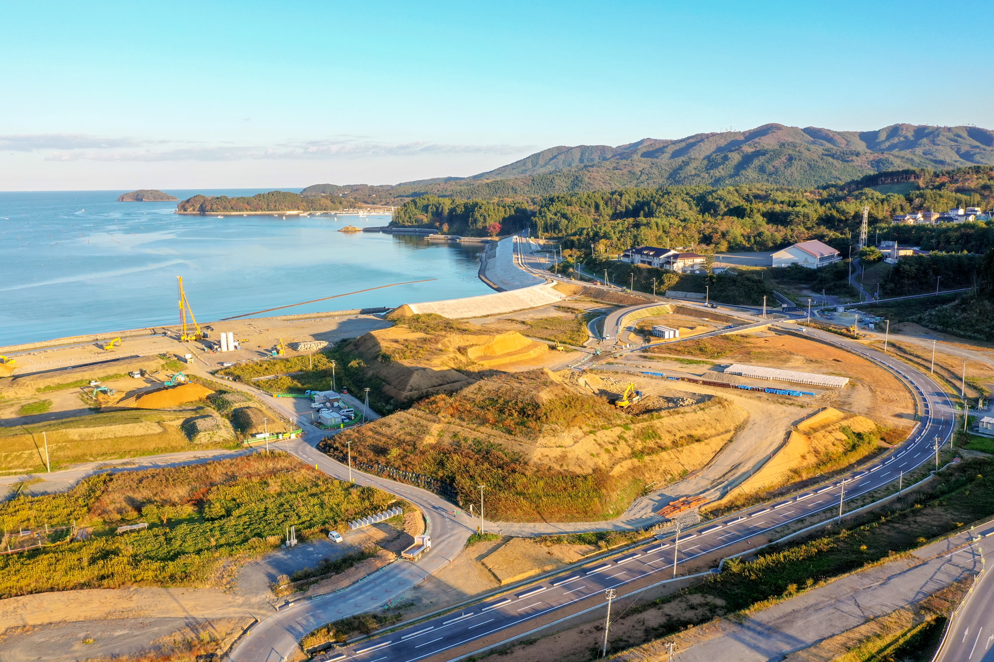 海に面した防潮堤の前で盛り土や工事が行われており、曲線を描く道路や重機が動いている開発地域の写真