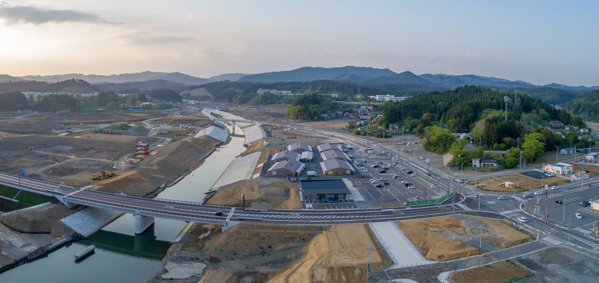 新しく建設された橋が川を横断し、その周囲に道路やインフラが整備され、工事現場や重機が点在する開発中の地域を上空から捉え、周囲には建物や駐車場、緑地が広がり、背景にはなだらかな丘と森林が見える晴天の風景のパノラマ写真