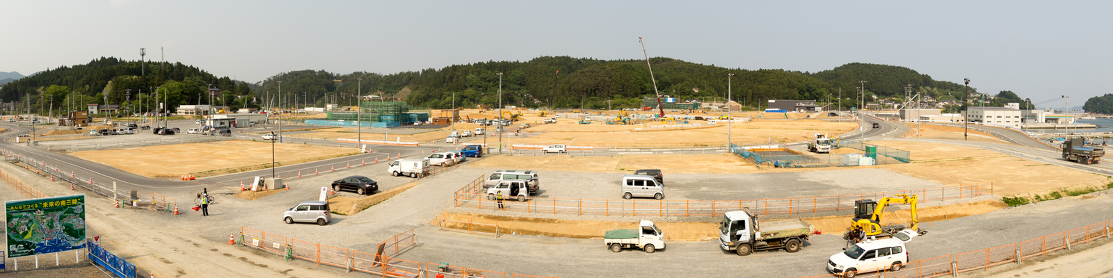 広い工事現場に複数のトラックやバン、建設機械が点在し、作業員が動いている様子が見られ、周囲は仮設フェンスで囲まれ、左側には工事に関する情報や地図が表示された案内板があり、背景には木々に覆われた丘が広がっているパノラマ写真