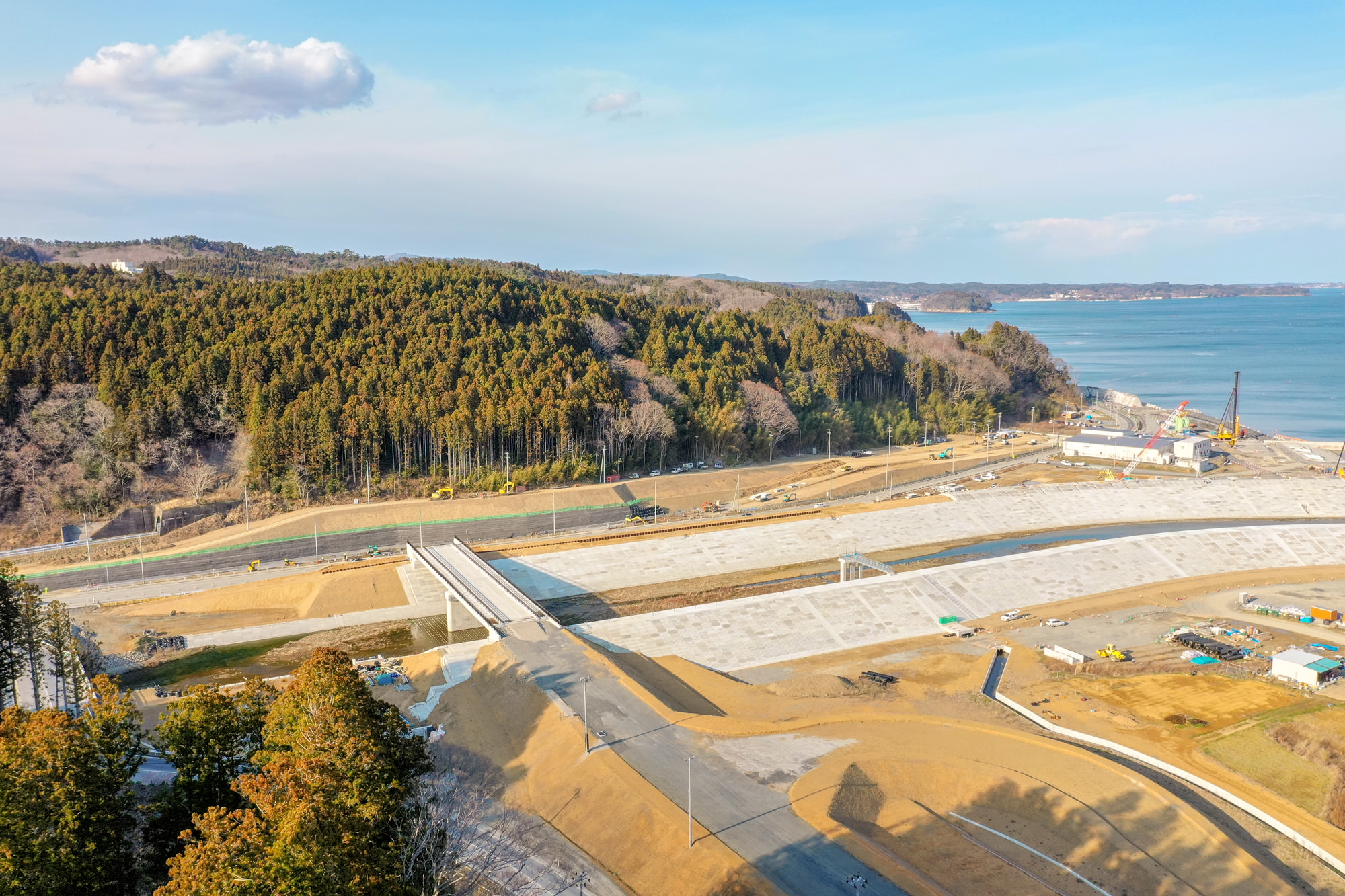 海に面した地域で新しく整備された堤防のようなインフラ構造物に橋が架かり、周囲には建設機材や資材が並び、背景には森林に覆われた丘と海が広がる沿岸防災施設の写真