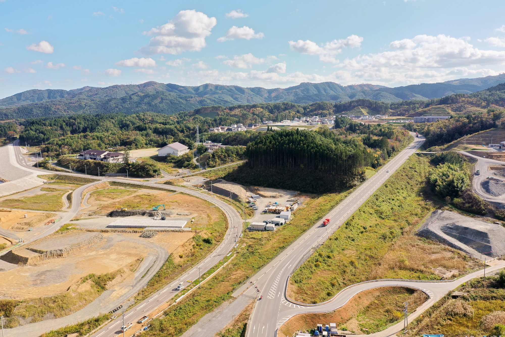 山間に延びる広い道路と湾岸に向かう工事中の道が交差し、奥には山並みと住宅地が見える、内陸と海岸の接点をとらえた復興地域の写真