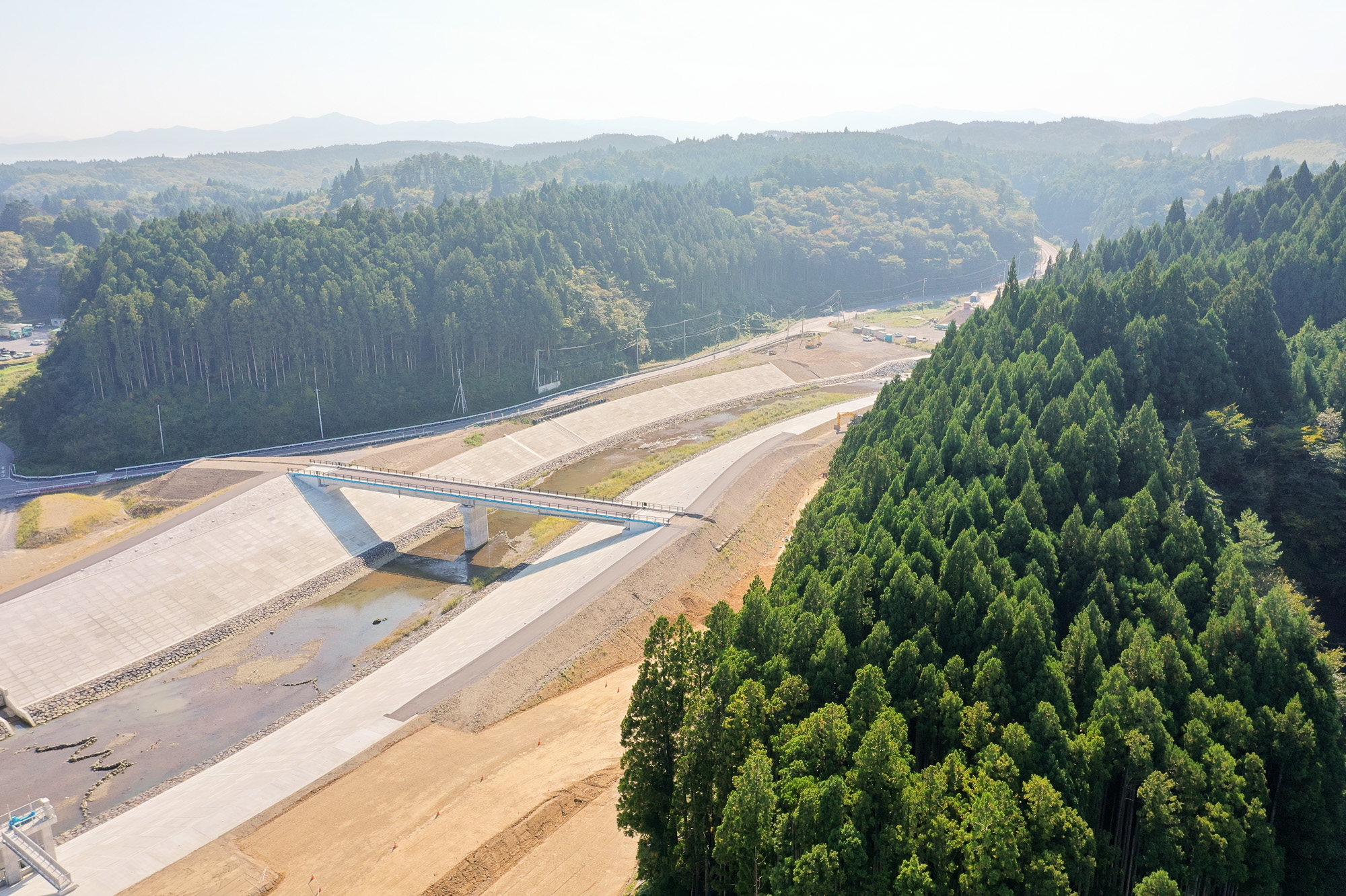 防潮堤の上を横断する橋と、それに沿って山間部へ続く道路、右側には濃い緑の杉林が広がっている写真
