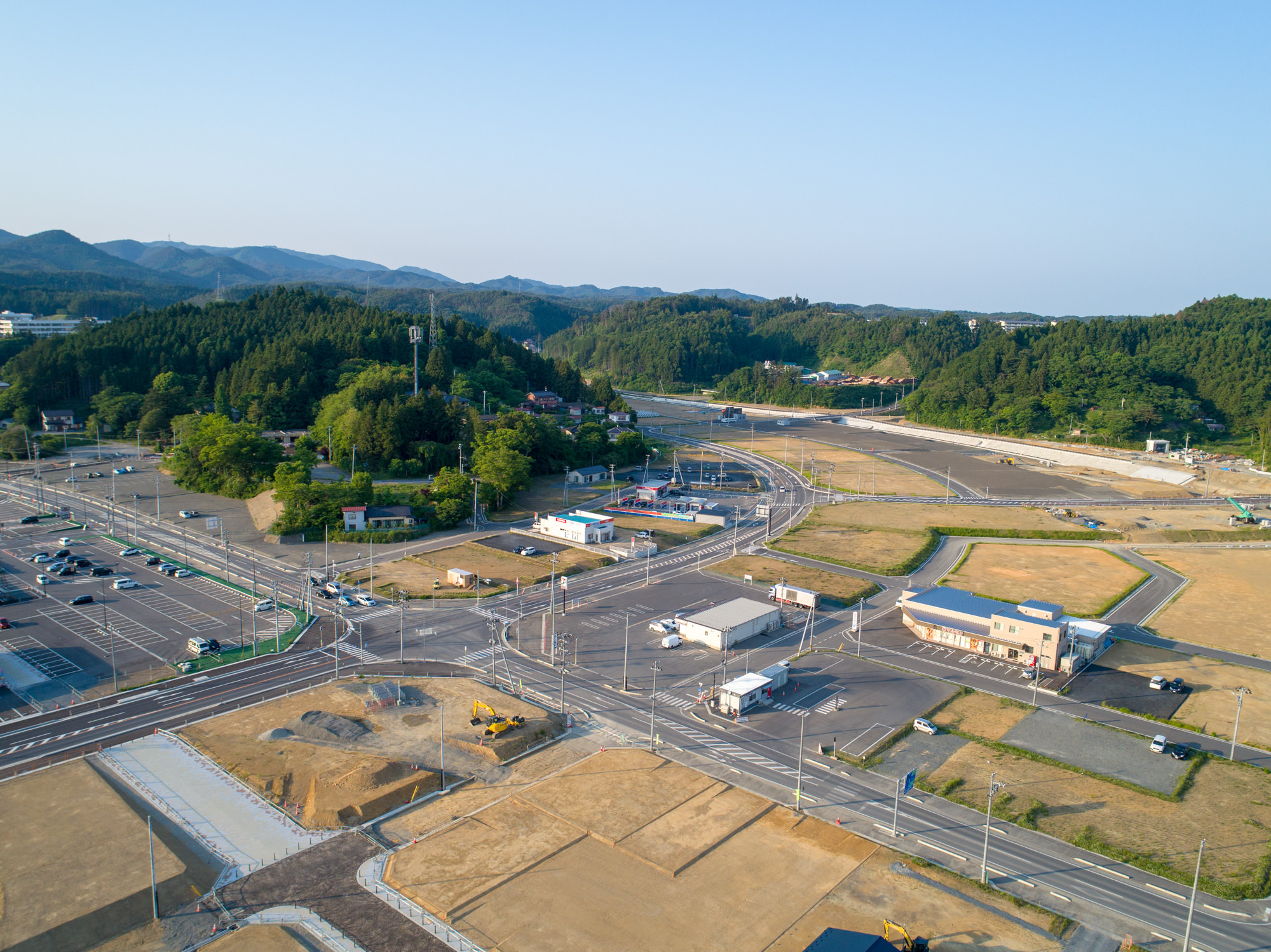 開発途中の半都市型地域を上空から撮影したもので、建物や道路、駐車場が点在し、一部の土地が整地されており、車両や建設機械が見られ、背景には緑の丘と森林が広がる風景の写真