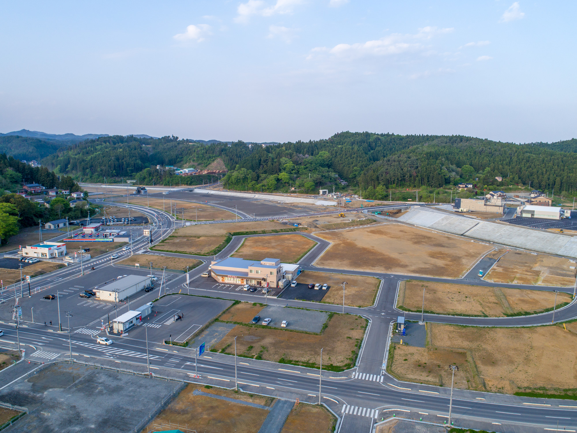 複数の道路や交差点、建物が広がる開発済みの地域を上空から捉えた写真で、商業施設や住宅地と思われる区域に加え、空き地も点在し、背景には緑の丘や木々が広がる郊外または田舎の風景が、晴れた空の下に撮影された写真