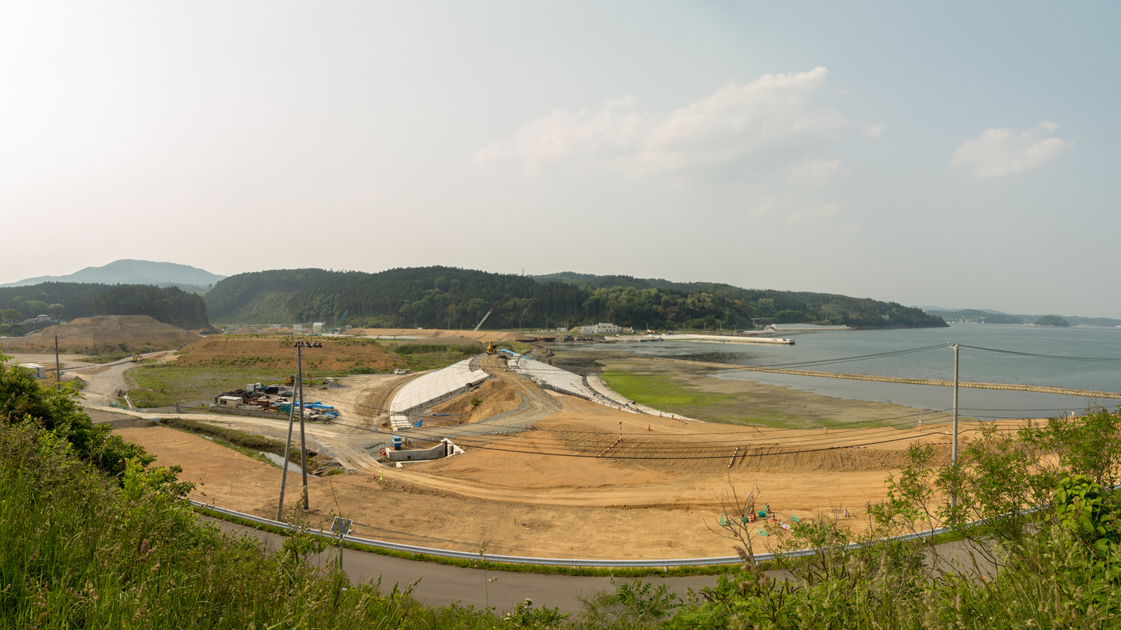 青い海に面した海岸沿いの広い工事現場で、重機が作業を行っており、周囲には舗装された道路や建設資材が整然と配置され、背景には水平線が広がる様子の写真