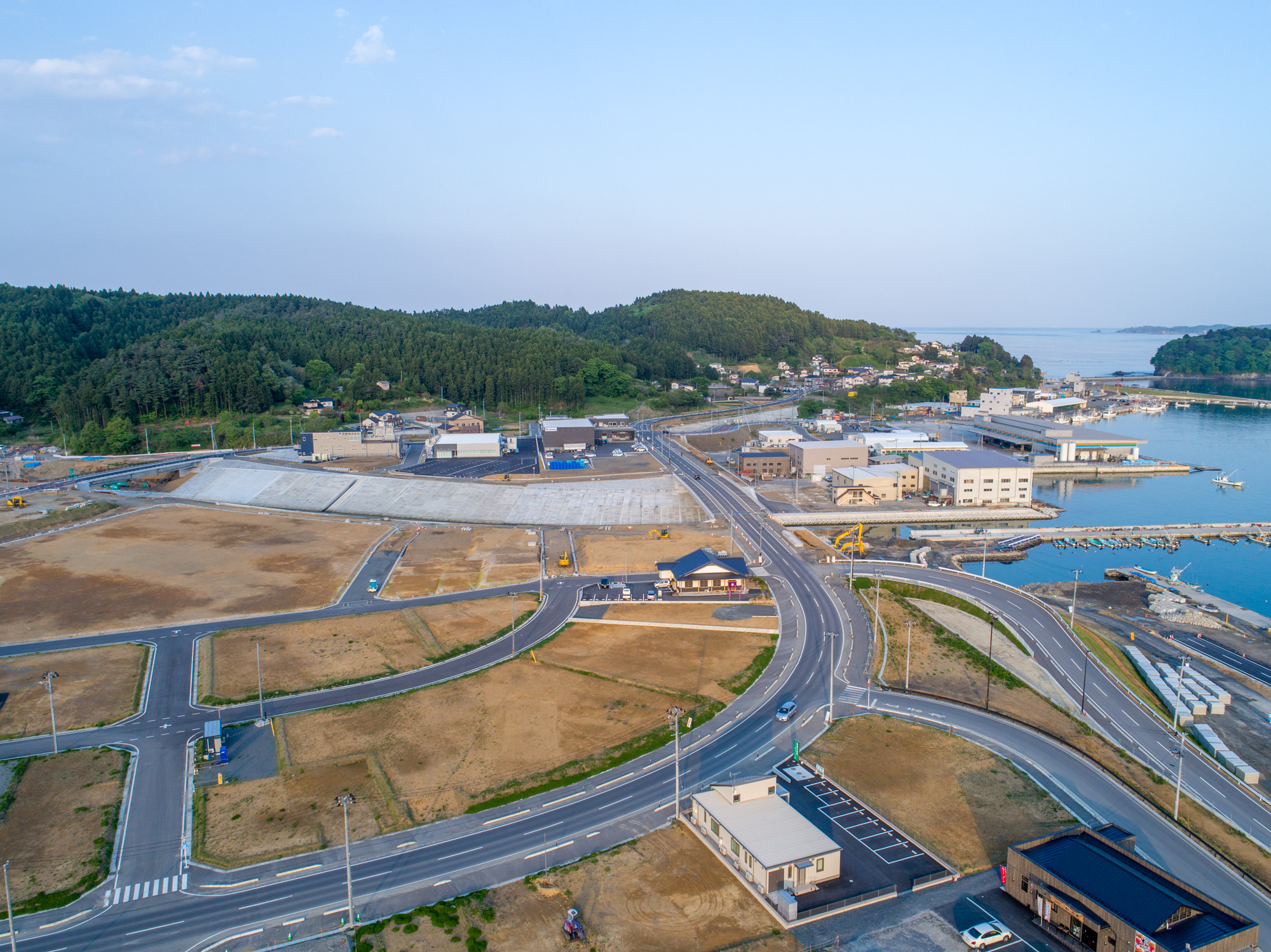 右側に港と船が見える海沿いの町を上空から捉えた写真で、建物や道路、空き地が混在し、道路は町の各所を結びながら海岸へと伸び、背景には緑豊かな丘や森林が広がる、静かな沿岸地域の風景の写真
