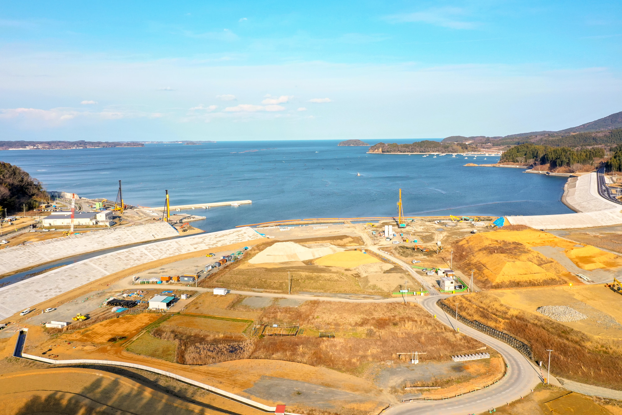 海に面した工事現場で、クレーンなどの重機が稼働し、整地された土地や構造物が点在し、背景には陸地が広がり、晴天のもとで作業が進められている様子を上空から捉えた沿岸工事の写真