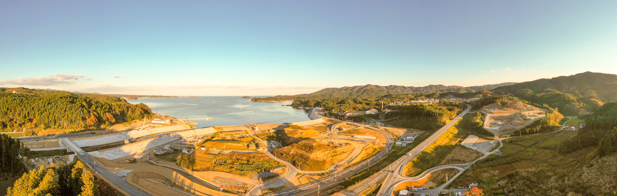 海に面した湾と山に囲まれた地域がパノラマで写っており、造成工事が広範囲に行われている様子が見える風景の写真