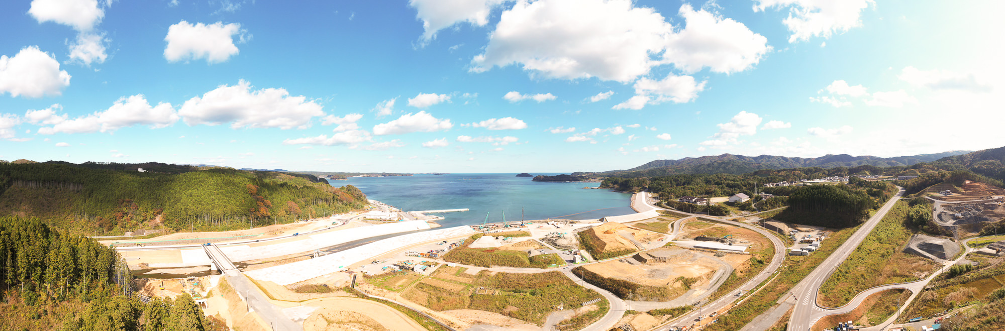 広い湾に面した海岸沿いに堤防や護岸工事が進められており、奥には森と山並みが続き、空は晴れて白い雲が浮かぶ青空の下での復興工事全体を見渡せる写真