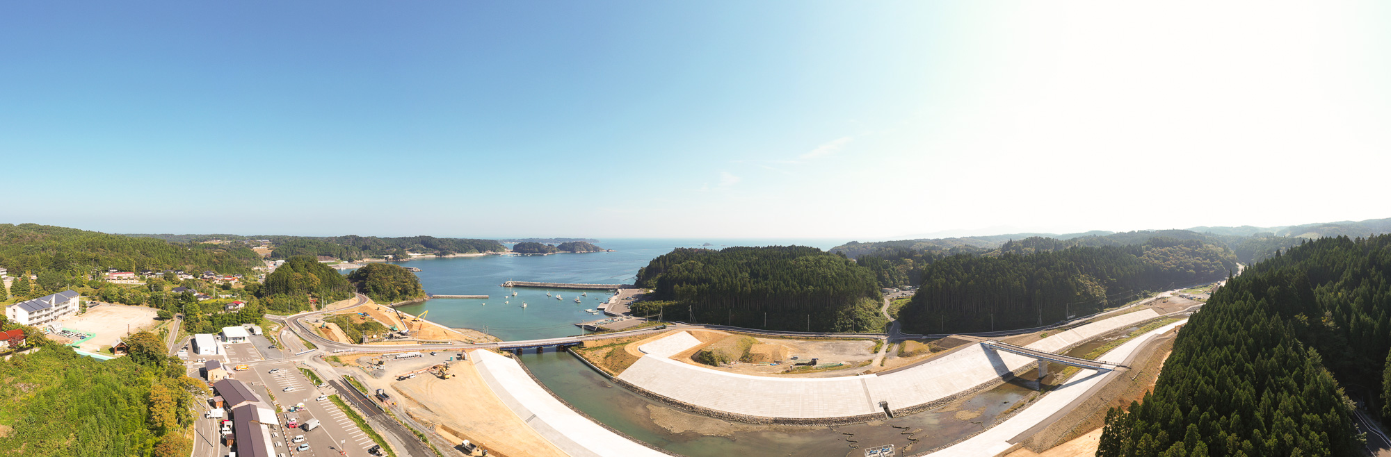山と海に囲まれた小さな港町を高い位置から広角で撮影しており、港、川、防潮堤、町の建物、森、道路が一望できる写真
