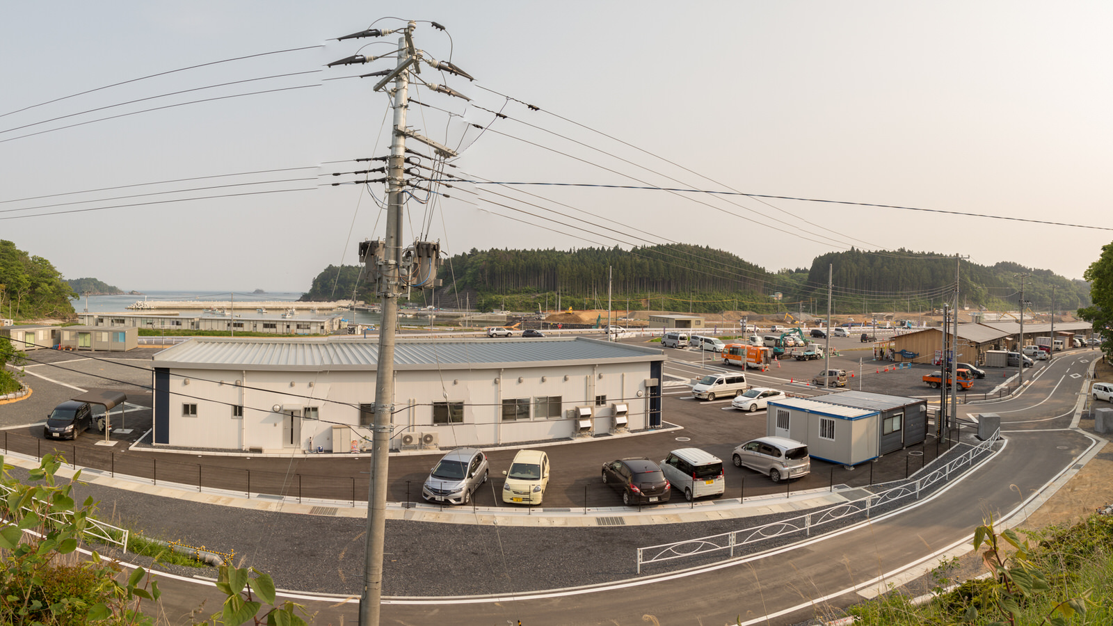 白い外壁と灰色の屋根を持つ平屋の建物を中心に、周囲に複数の車やトラックが駐車されている小規模な商業エリアで、電柱と電線が張り巡らされ、背景には緑の木々に覆われた丘と複数の建物が見え、空は雲のない晴天の風景の写真