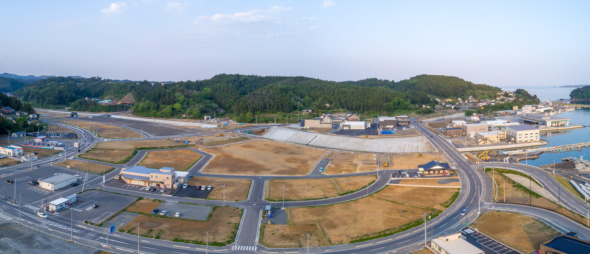 湾岸沿いに広がる町を上空から捉えた写真で、曲線や直線の道路が交差し、建物や空き地が点在し、道路上には車両も見られ、背景には緑豊かな丘や森林が広がり、右側には海または大きな川が見える、晴天の風景のパノラマ写真