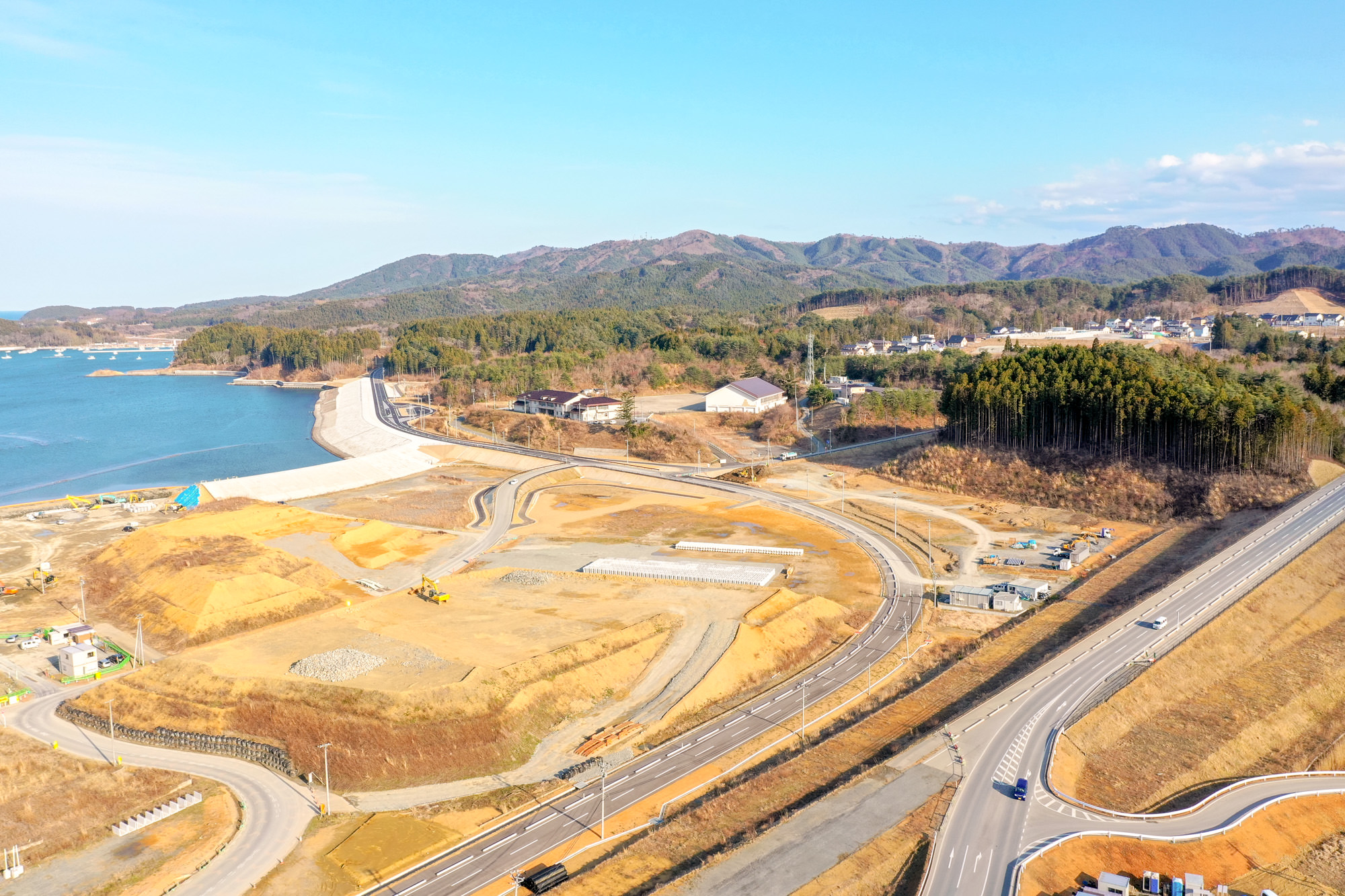 左側に海岸線が広がり、曲がりくねった道路や通路が各所へと続き、周囲には工事現場や森林に覆われた丘や山々が見える、自然と開発が混在する沿岸地域を上空から捉えた海辺の造成地の写真