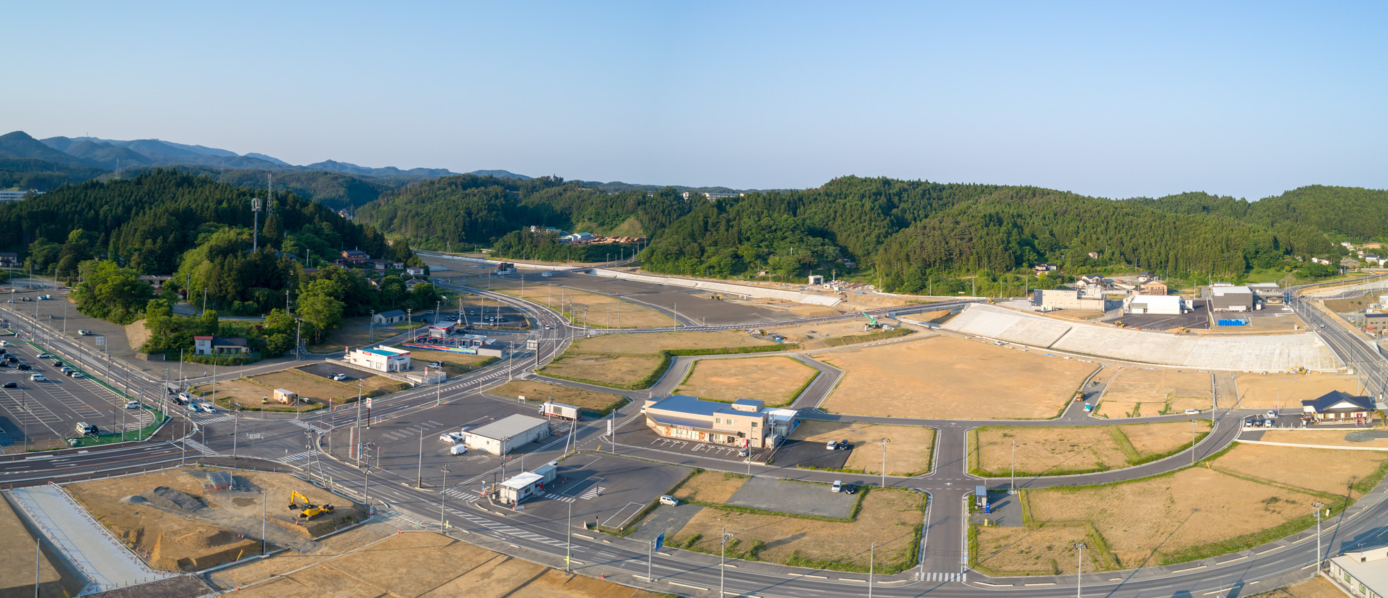 半都市型地域を上空から撮影したもので、建物や道路、駐車場が点在し、周囲には緑の丘や森林が広がり、雲のない青空が見える風景のパノラマ写真