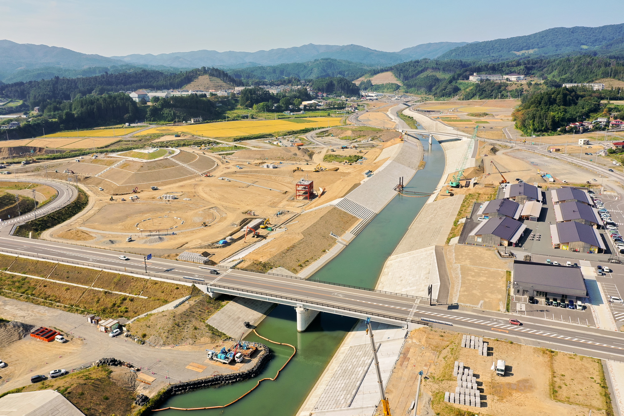 整備中の護岸に囲まれた川が中央に流れ、その上を道路橋が通り、周囲では造成工事が進められている様子が見える風景の写真