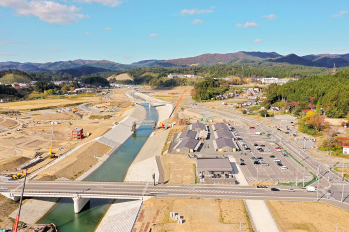 広い谷あいを縫うように川が整備され、手前に橋がかかっており、川の右側には建物と広い駐車場、奥には山並みが広がる風景の写真