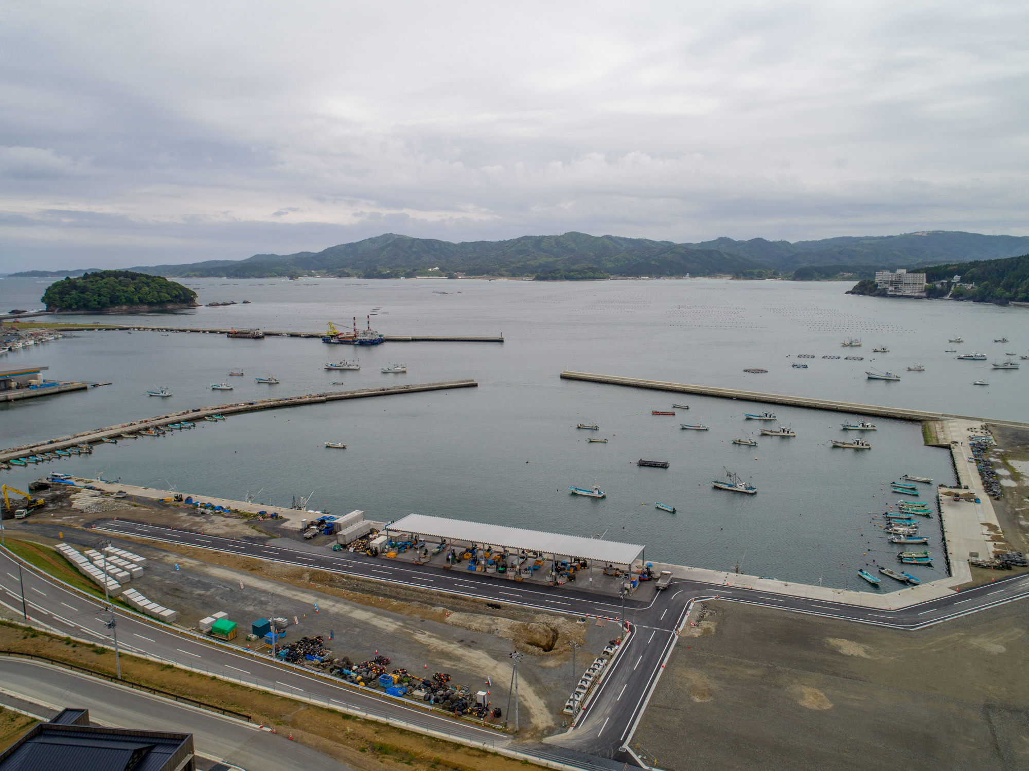 防波堤に囲まれた港に複数の船が停泊している海岸地域を上空から捉えた写真で、海岸沿いには建物や道路が並び、背景には丘や山々が広がり、空には雲がかかっている風景の写真