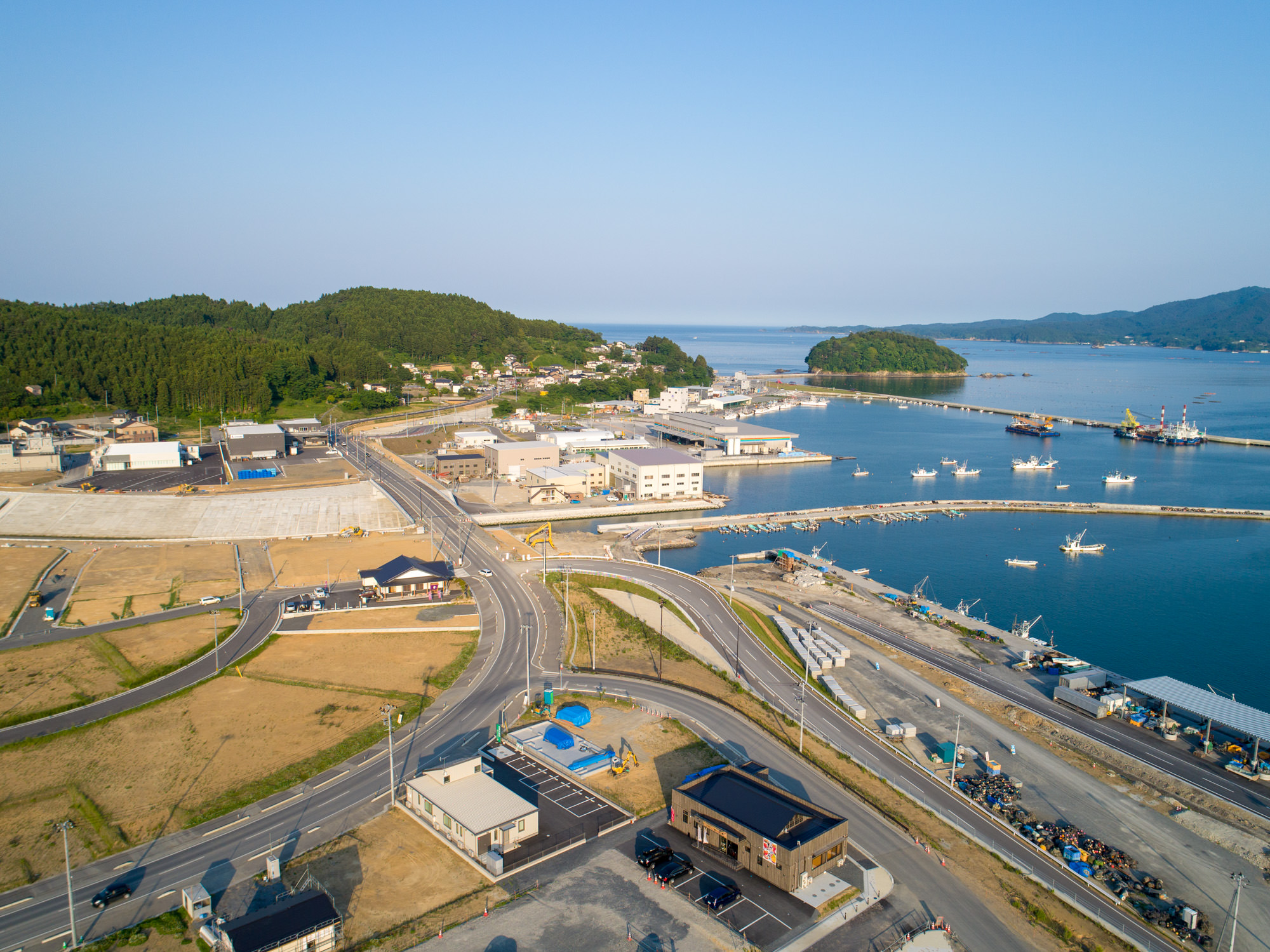 港を中心とした海沿いの町を上空から撮影したもので、穏やかな水面に複数の船が停泊し、周囲には建物や道路、背景には緑の丘と防波堤が見える風景の写真