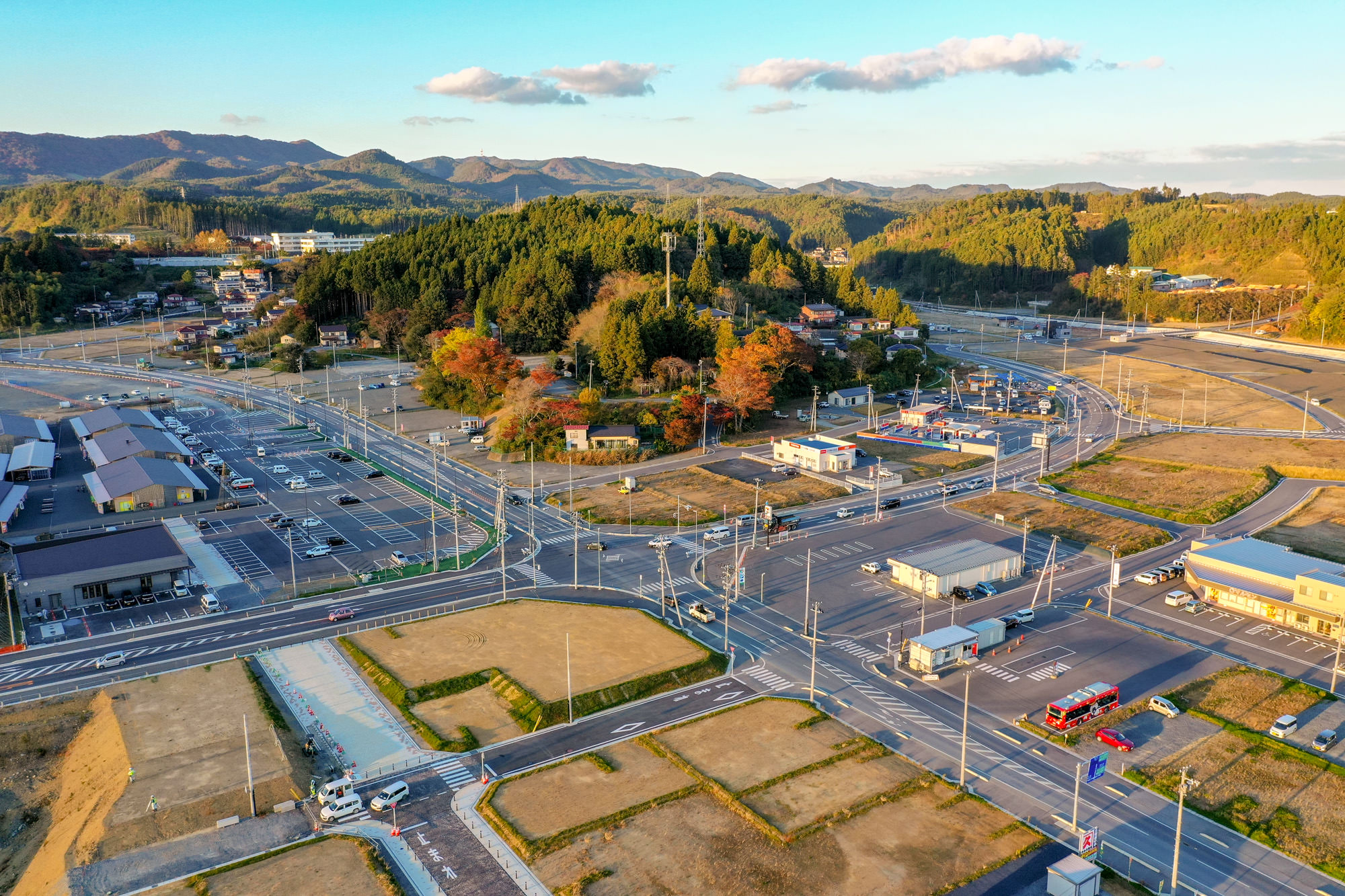 整備された交差点と広い駐車場、建物群、紅葉した木々に囲まれた小高い森が広がる地域の町並みを山を背景に上空から撮影した写真