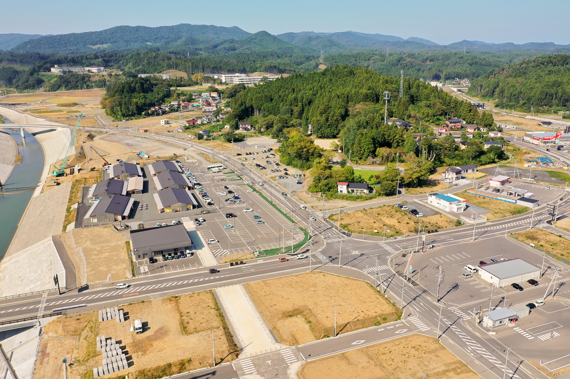 川沿いに立ち並ぶ商業施設群と広い駐車場、その奥に住宅と森林が広がっている様子を上空からとらえた風景の写真