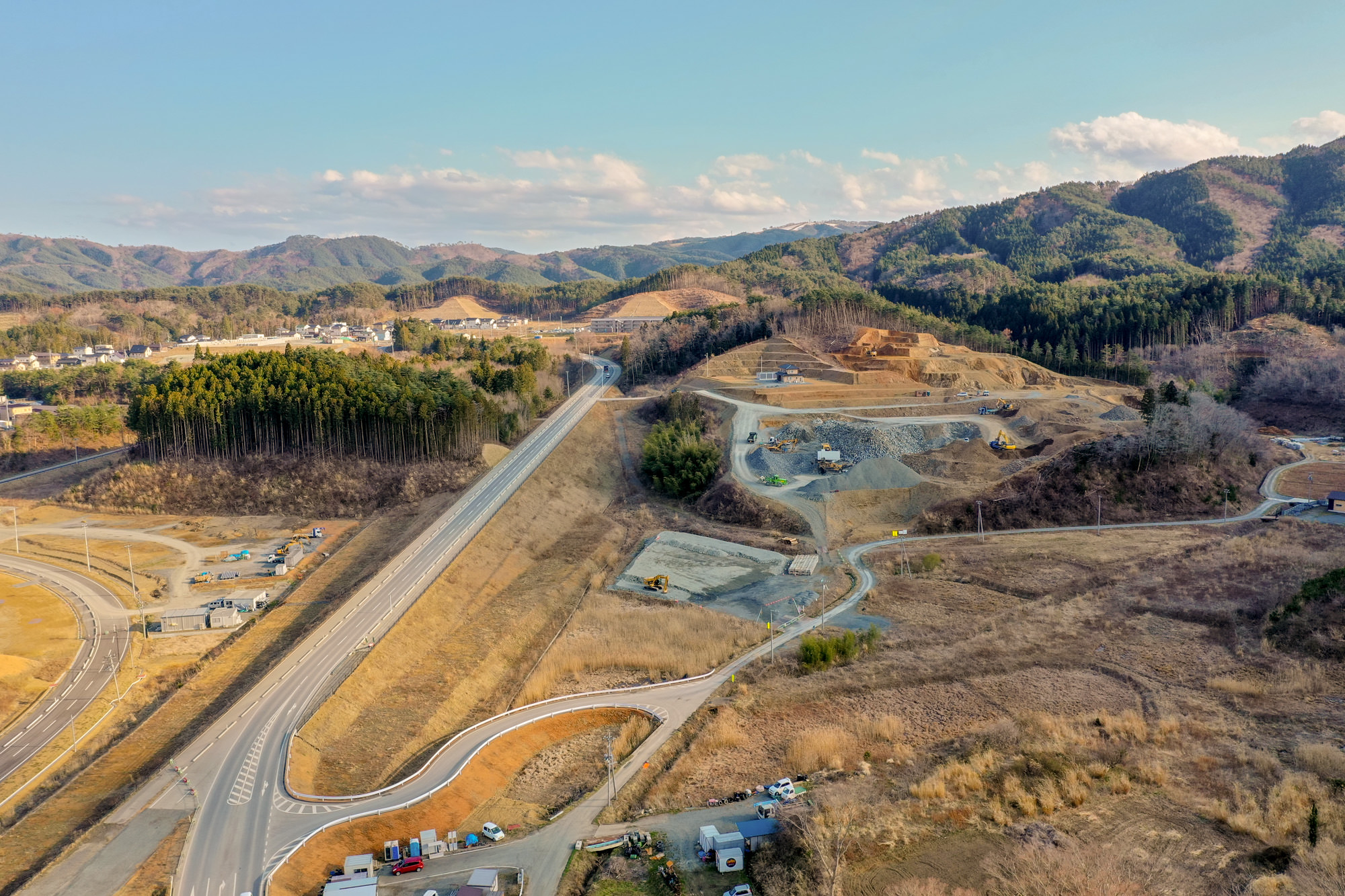 森林に覆われた丘陵地帯を貫く高速道路が広がり、複数の道路や建設中の区画、点在する建物が見え、背景には山々と雲が浮かぶ空が広がる交通インフラ整備中の地域を上空から捉えた山間部の道路開発の写真