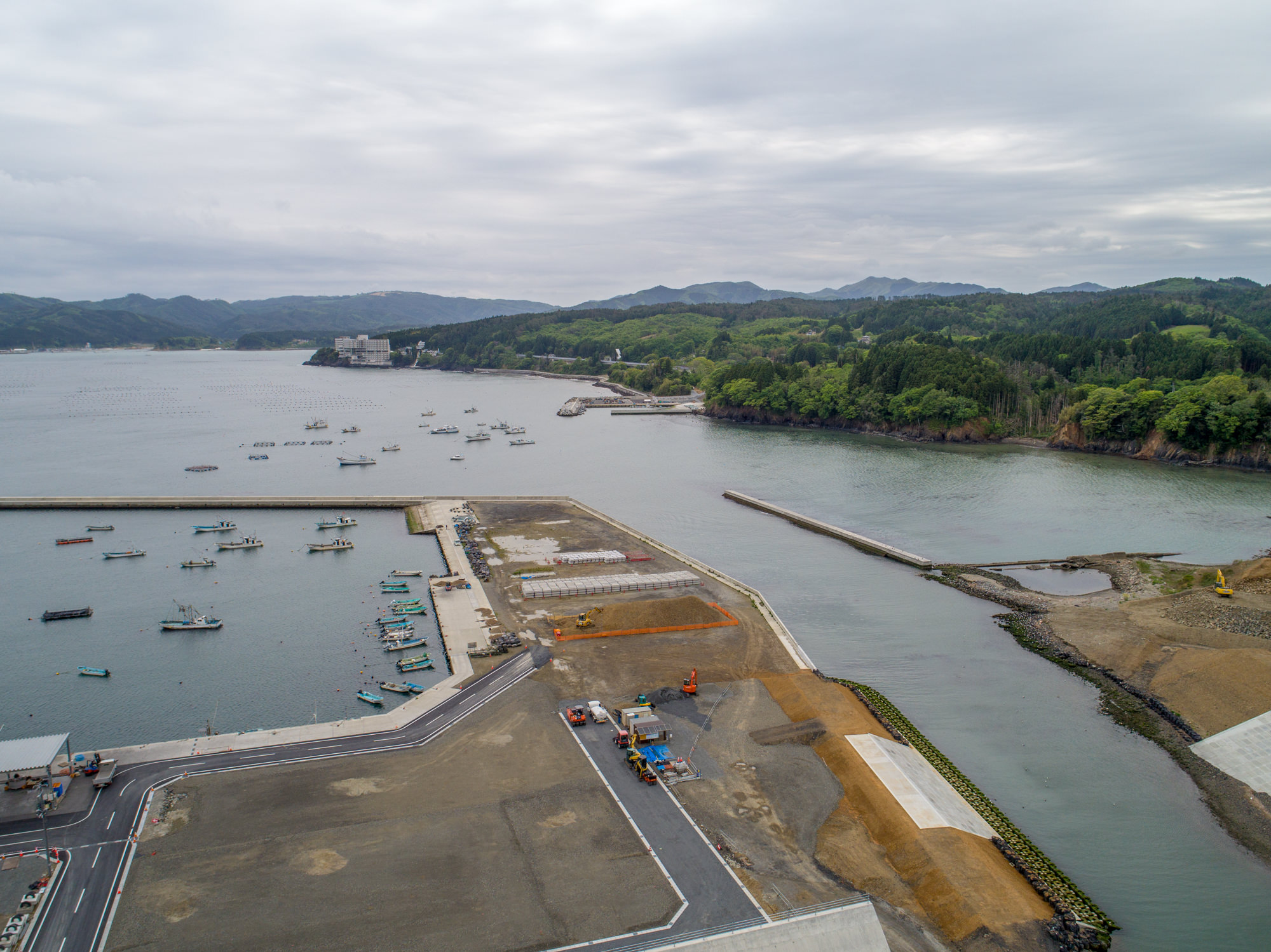 防波堤に囲まれた港に複数の船が停泊し、隣接する陸地では建設機材や資材が見られる工事中の様子が広がり、背景には緑豊かな丘や山々、海岸近くには建物が点在し、曇り空の下で撮影された海岸地域の上空からの風景の写真