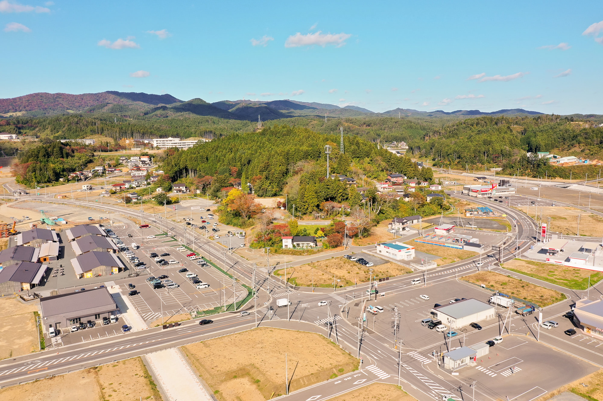 山のふもとに広がる住宅街と学校施設、左側には整備された商業施設と駐車場があり、その手前には交差点と複数の車両が走る市街地の写真