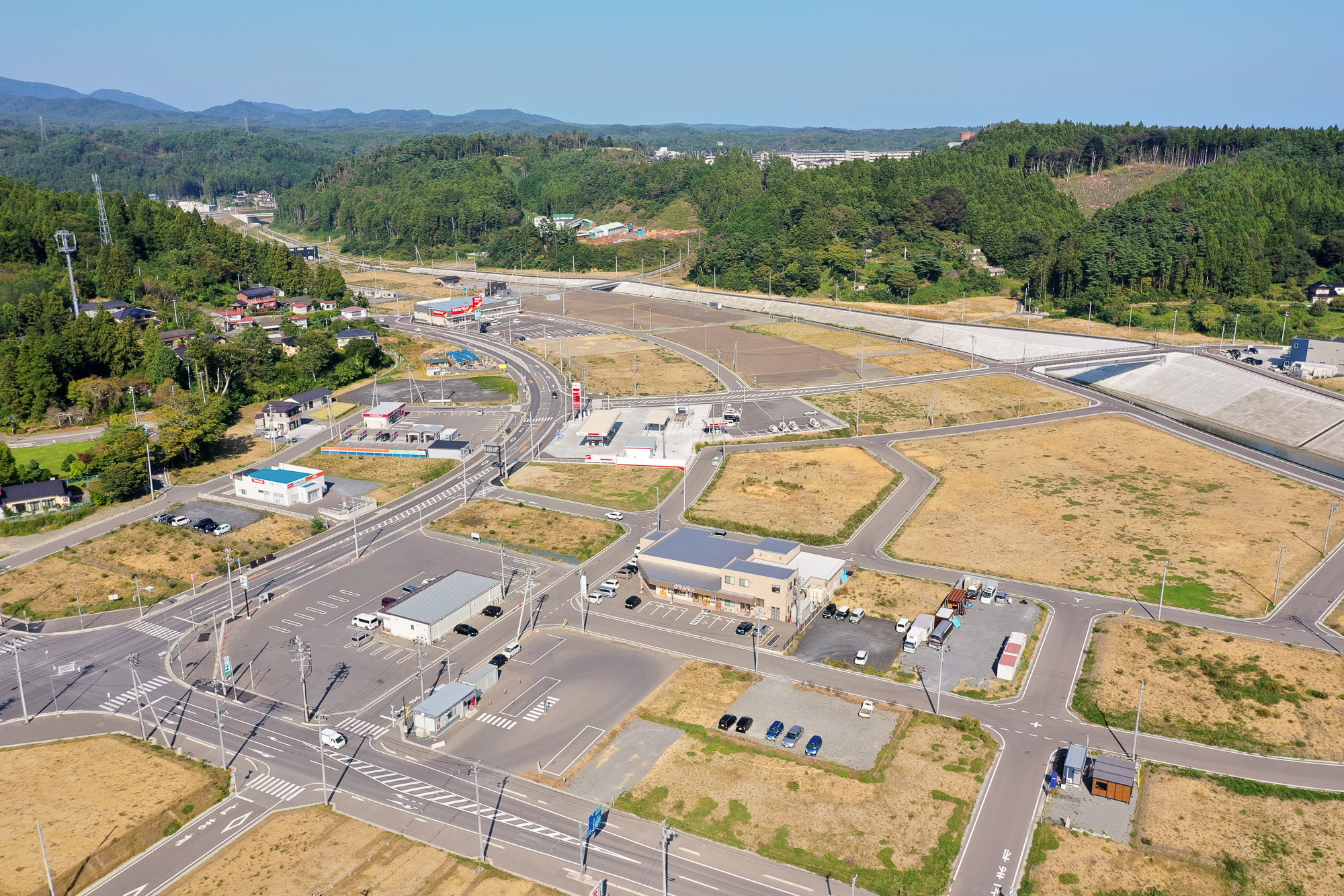 整備された道路網の交差点に囲まれた商業施設や駐車場が点在し、その奥に山間の住宅地が広がる様子の写真