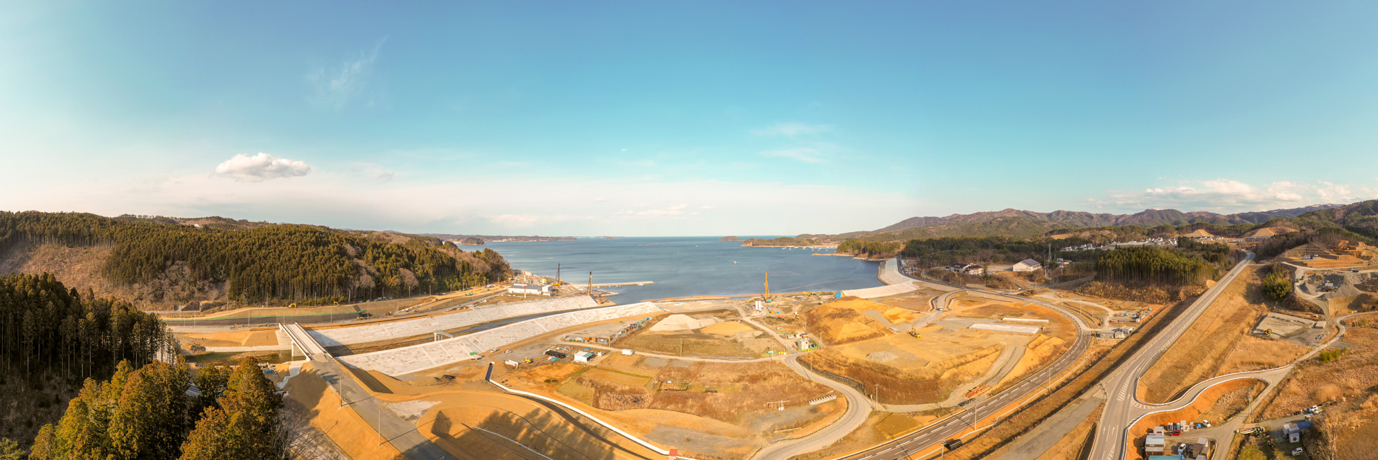 海を背景に、両側に広がる丘や森林に囲まれながら、手前には複数の工事現場や道路、インフラ整備が進む様子が見える自然と開発が共存する沿岸地域を上空から捉えた海辺の開発地の写真