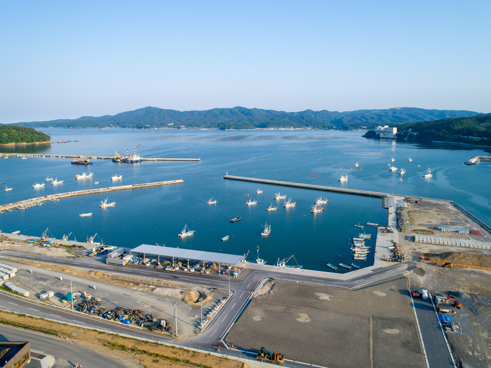 複数の船が停泊・浮かぶ港を上空から撮影したもので、防波堤に囲まれた港の周囲には建物や工事中のエリアがあり、背景には丘や山々、青空が広がる風景の写真