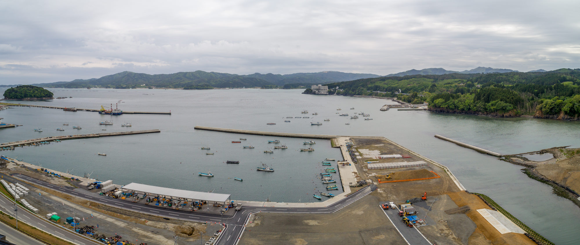 港に複数の船が停泊し、いくつもの防波堤が海に突き出している海岸地域をパノラマで捉えた写真で、海岸沿いには建物や道路が並び、背景には緑豊かな丘や山々が広がり、空には雲がかかっている風景の写真