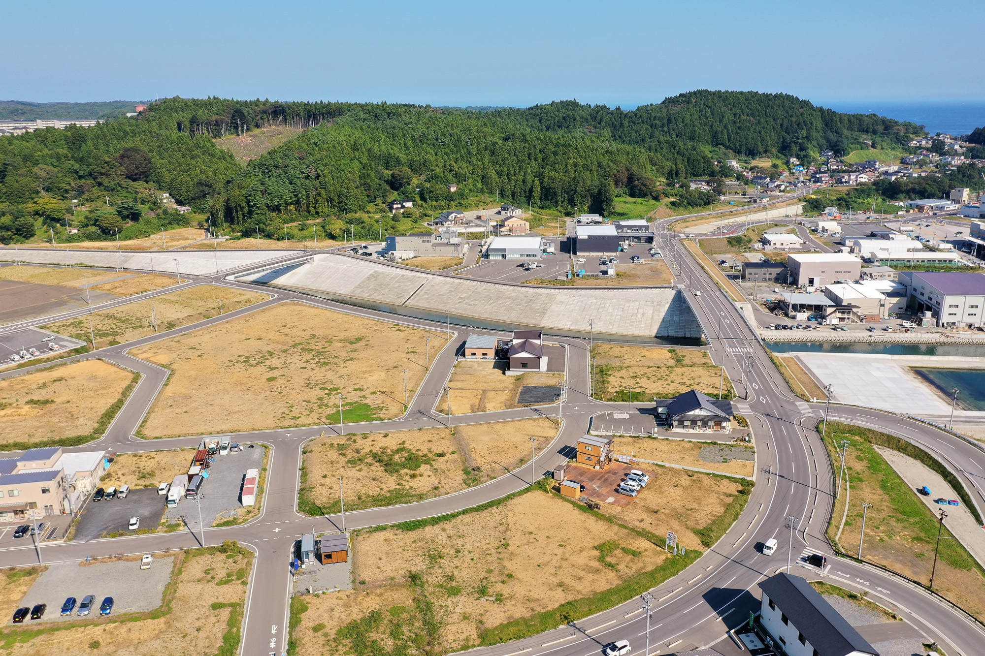 区画整備された土地に小さな建物や倉庫が点在し、奥にはコンクリート護岸と緑の山が見える風景の写真