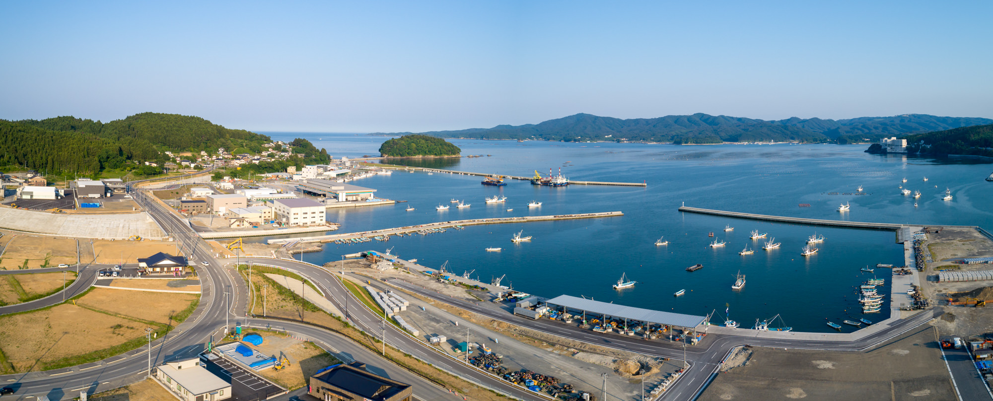 港町を上空から撮影したもので、港には多数の船が停泊し、いくつもの桟橋が海に伸びており、周囲には建物や道路、背景には緑の丘が広がり、青空が広がる晴天のパノラマ写真