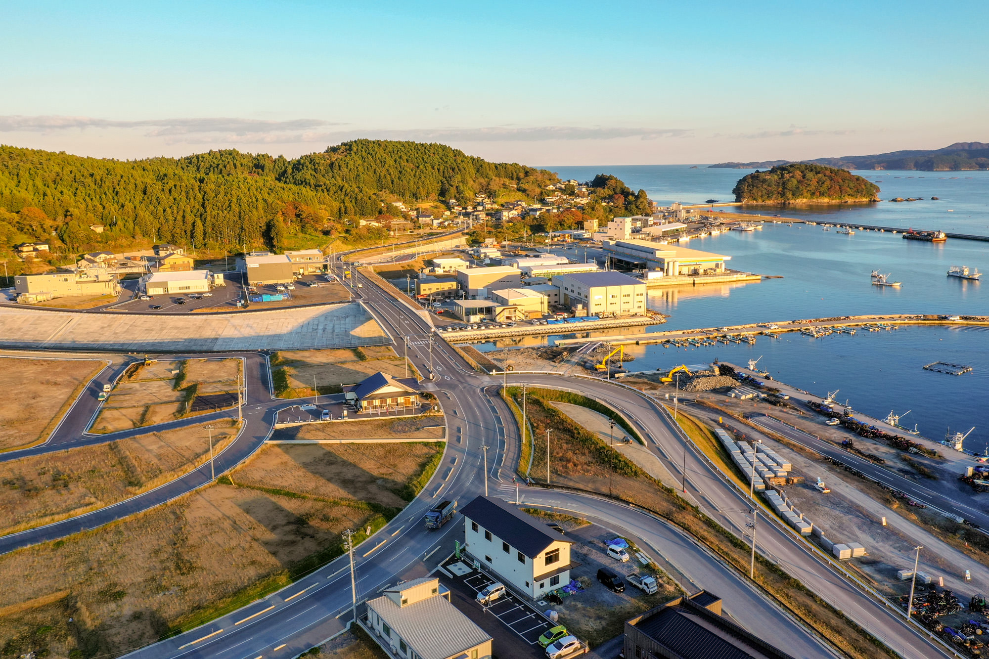 整備された道路が緩やかに曲がりながら港へ続き、周囲に建物や駐車場、そして島や漁港、海岸が見える海辺の町を撮影した写真