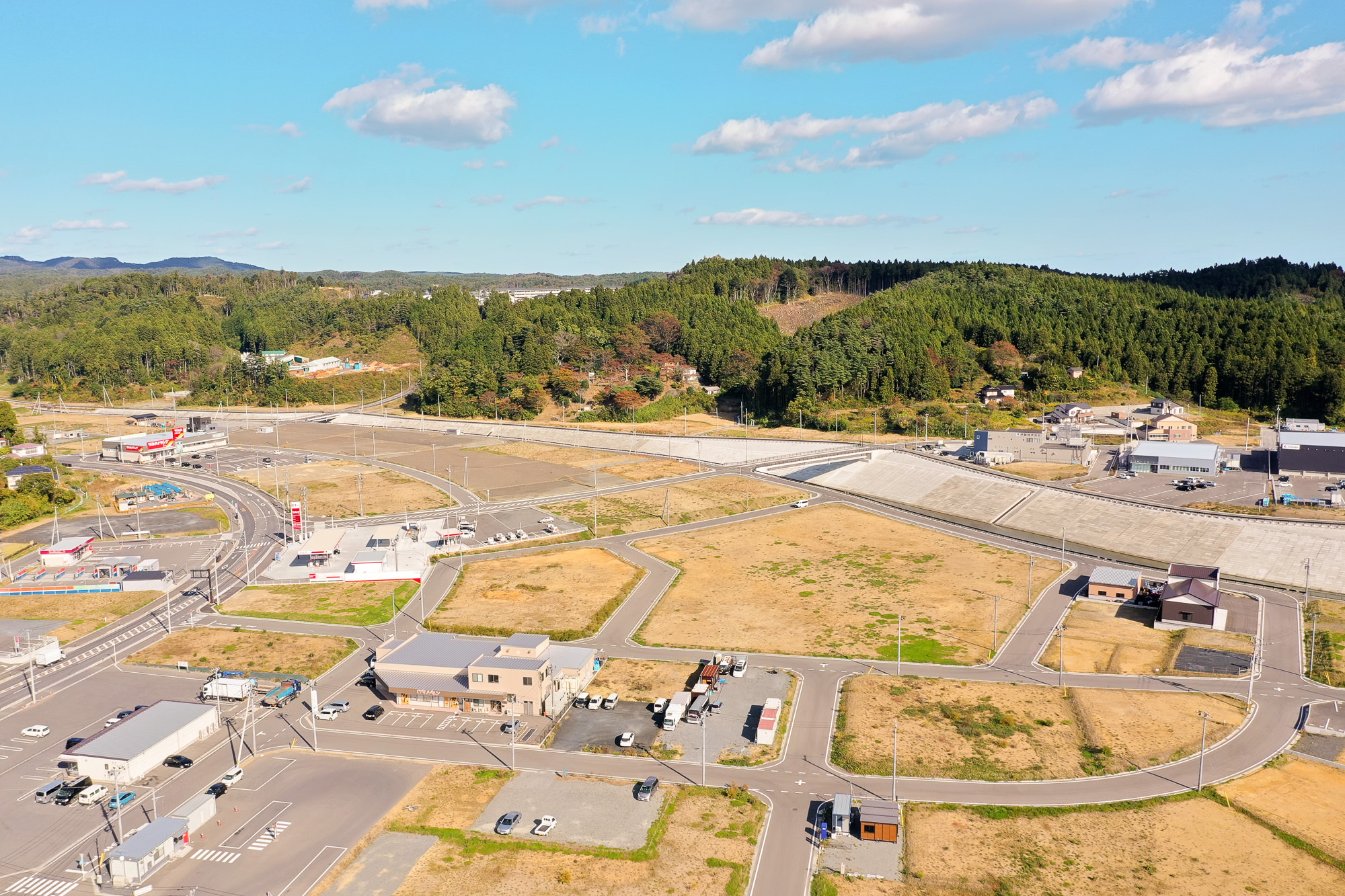 道路網と区画整理が進んだ広大な土地の中に点在する建物や商業施設があり、右奥には高く積まれた堤防が見える郊外の復興造成地の写真