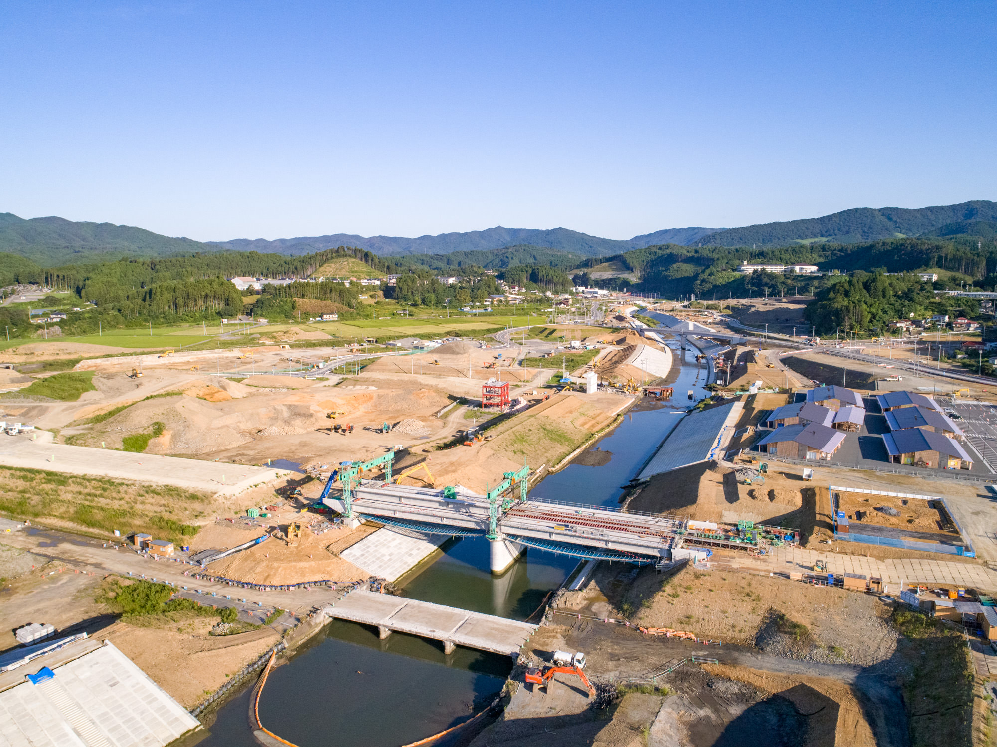 中央に川が流れ、その上に複数の橋が建設中である広大な建設現場を上空から撮影したもので、周囲には工事用の機材や車両、資材が点在し、背景には緑の丘や森林が広がり、青空が広がる建設現場の写真