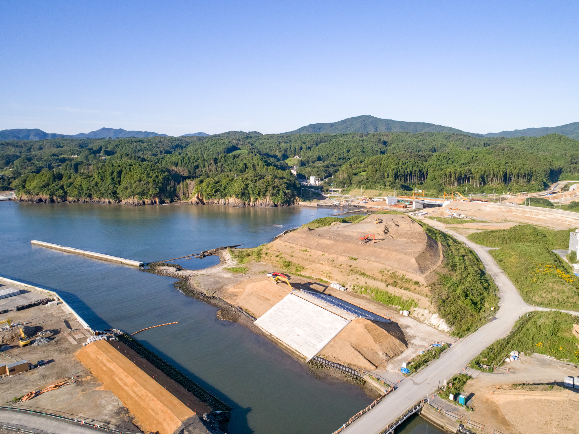 海辺にある建設現場を上空から撮影したもので、複数の構造物や土木工事が進行中であり、隣接する水域のそばに道路や堤防、大きな傾斜のあるコンクリート構造物が見られ、背景には緑の丘や森林が広がっている沿岸部の建設現場の写真