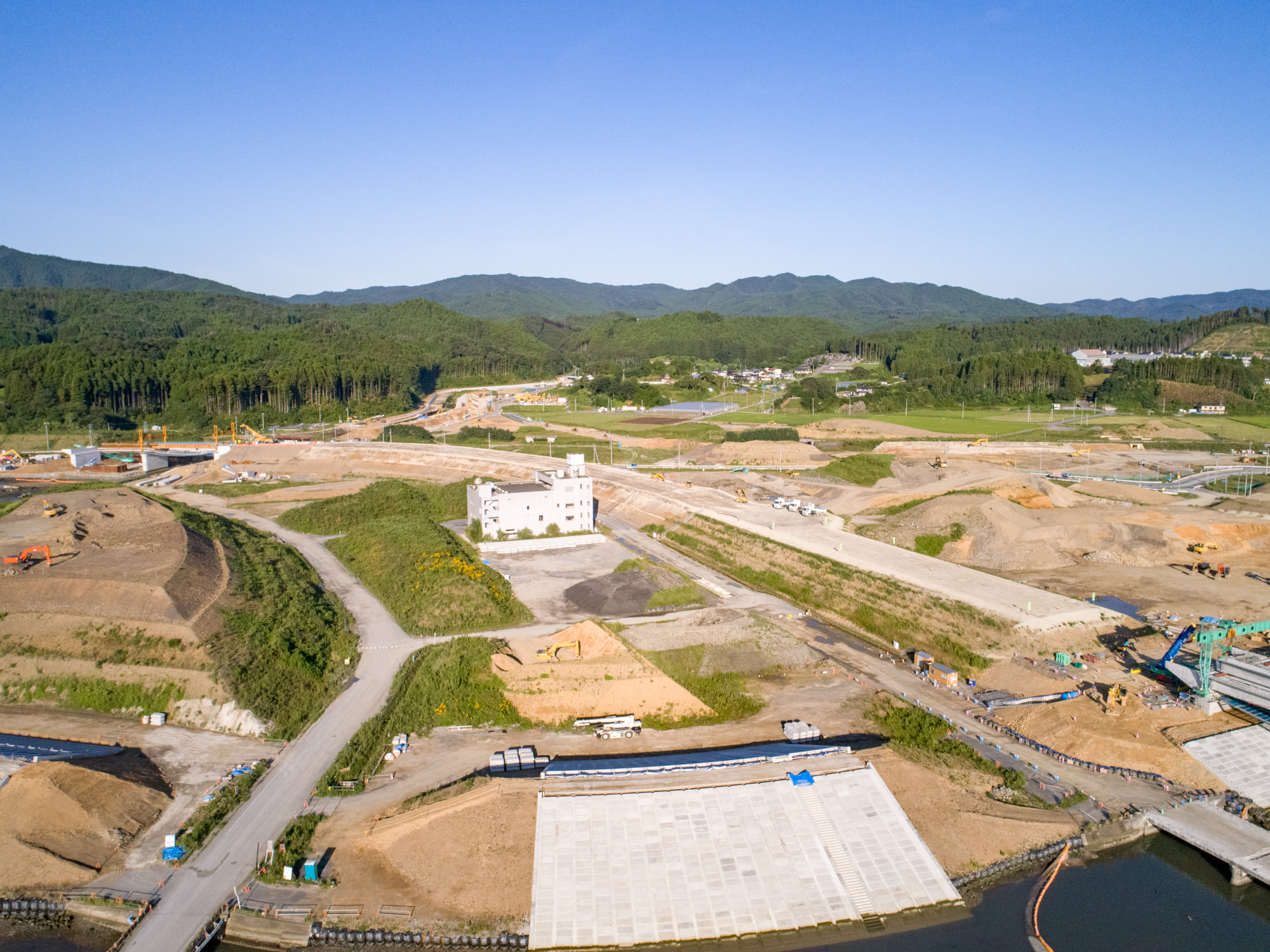 志津川地区にある建設現場を上空から撮影したもので、複数の未舗装の道路や小道が走り、建設機材が点在し、中央付近には建設途中の建物があり、周囲には丘や森林が広がり、背景には山々がそびえ、青空が広がる建設現場の写真
