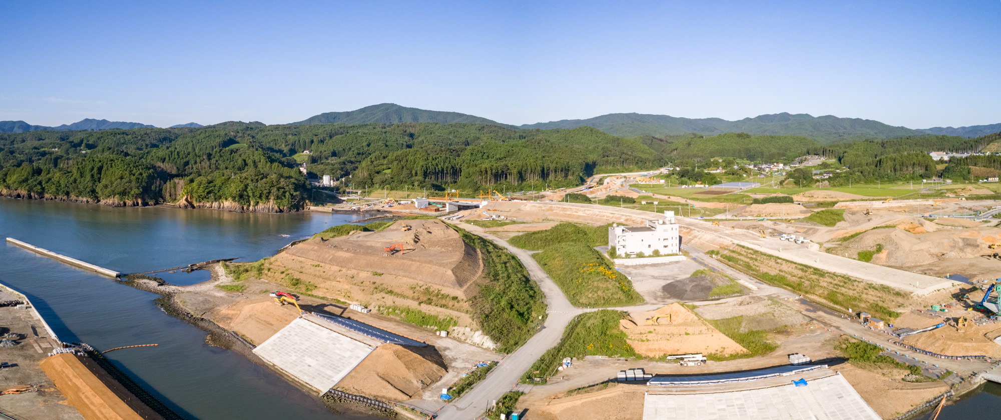 川沿いにある建設現場をパノラマで上空から撮影したもので、複数の大きな土の山、建設機材、建設途中の構造物があり、中央右には白い建物が見え、周囲には緑の丘や森林が広がり、背景には山々がそびえ、青空が広がる建設現場の写真