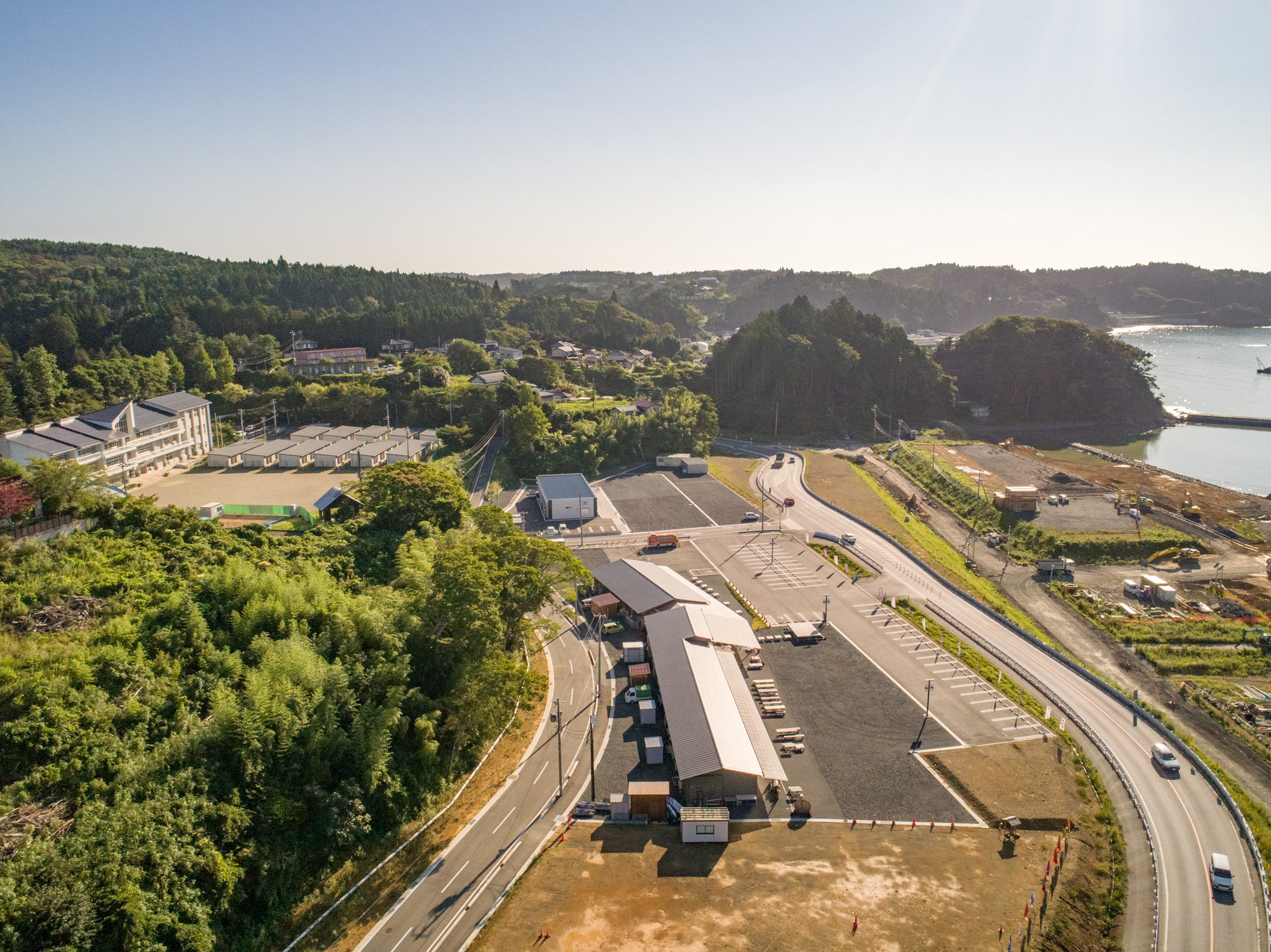 自然と開発された要素が混在する沿岸地域を上空から撮影したもので、建物、道路、駐車場、緑地が見られ、背景には海岸線と水域が広がっている工事現場の写真