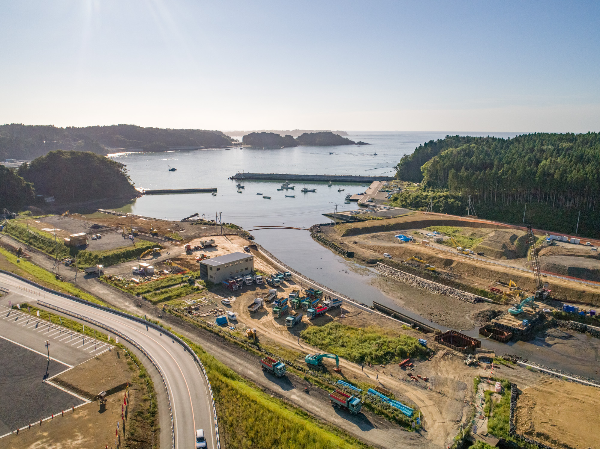 海辺にある建設現場を上空から撮影したもので、複数の工事用車両や機材が点在し、現場内にはいくつかの建物や構造物があり、港には船が浮かび、周囲には緑地や木々が広がり、敷地の縁にはカーブを描く道路が通っている沿岸部の建設現場の写真