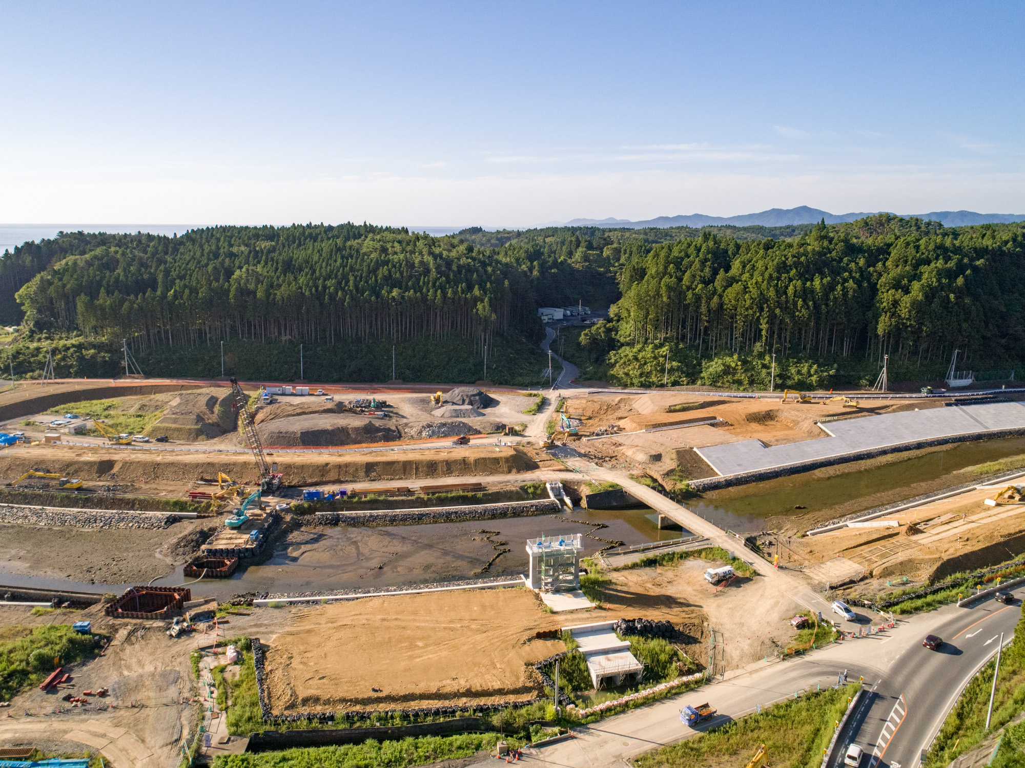複数の工事区画で土木機材や資材が配置され、建設作業が進行中の建設現場を上空から撮影したもので、背景には森林が広がり、現場内には道路や小道が通じており、水域が敷地の一部を流れ、橋や渡り道が架けられている、丘陵地帯に位置する建設現場の写真