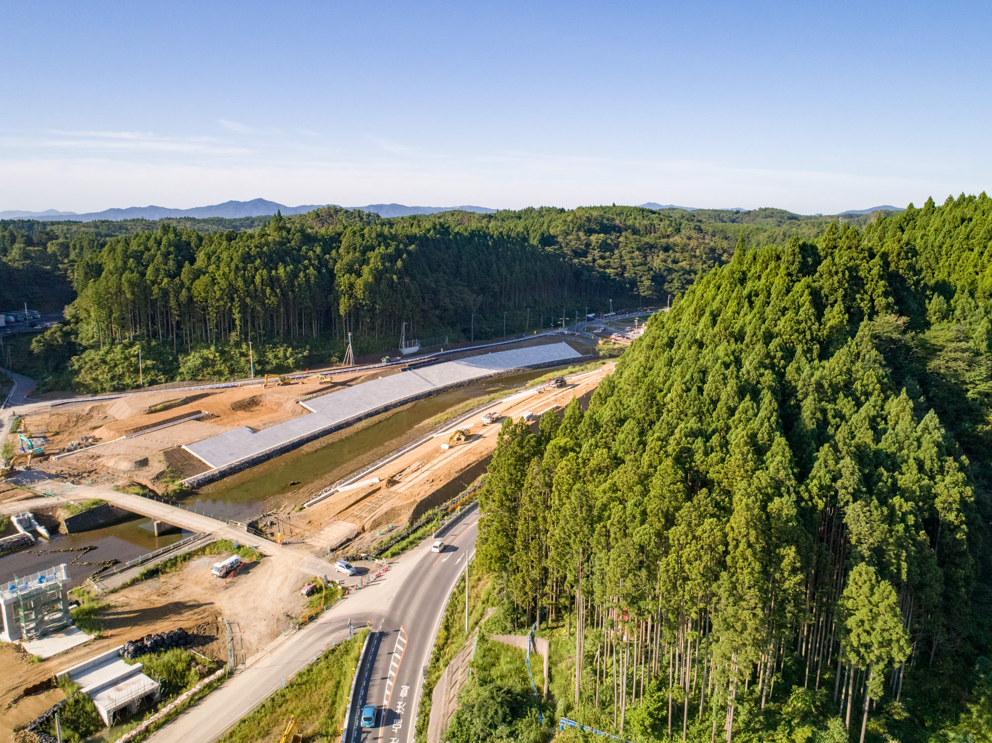 森林に囲まれた建設現場を上空から撮影したもので、手前には現場を囲むようにカーブした道路があり、背景には青空の下に広がる緑豊かな森林と丘が見える森林地帯の建設現場の写真