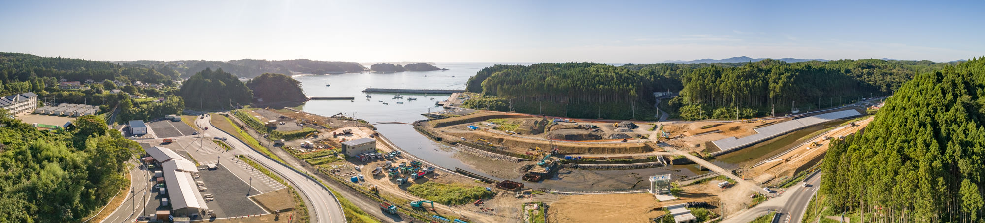 建設中の沿岸地域をパノラマで上空から撮影したもので、複数の工事現場に機械や設備、道路、建設中の建物が点在し、背景には船や島が浮かぶ水域が広がり、周囲には緑豊かな森林と丘が広がり、晴天のもとで撮影された建設現場の写真