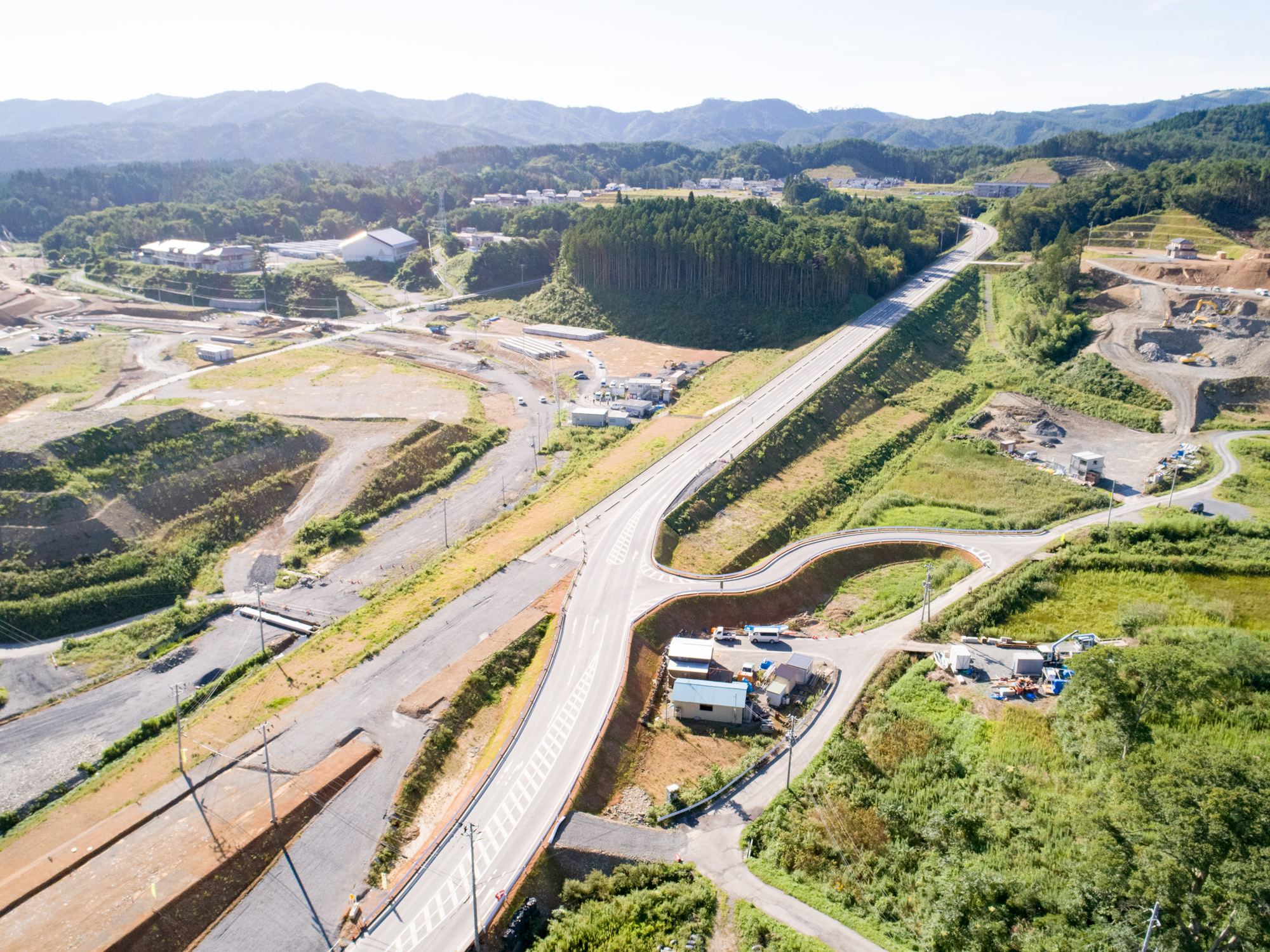 複数車線の新設道路がカーブしながら敷地を横断し、一部は盛土で高くなっており、周囲には建設現場や建物、緑地が点在し、背景には木々に覆われた丘や山々が広がる地域を上空から捉えた道路建設現場の写真