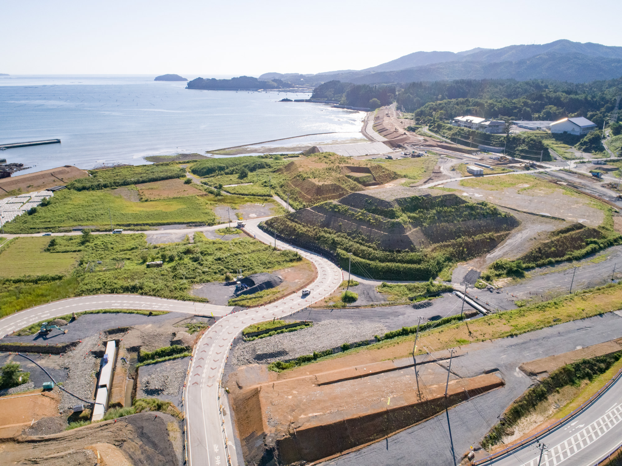 海を背景に、複数の曲がりくねった新設道路や段々に整備された土地、緑地、そして海岸近くに建物が点在する様子を、開発中または開発直後の沿岸地域として上空から捉えた造成地の写真