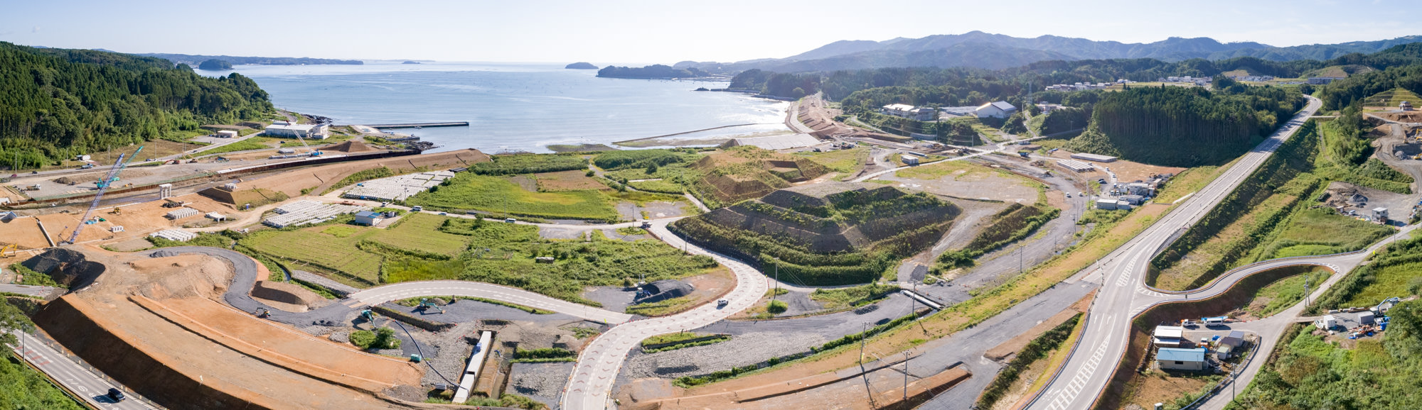 舗装・未舗装の道路が曲がりくねりながら広がり、整地された土地や建設機材が点在し、緑地や畑の丘陵地が混在する沿岸の開発地域を、遠くに海と延びる海岸線、周囲の森林や山々とともに上空から捉えた沿岸工事現場のパノラマ写真